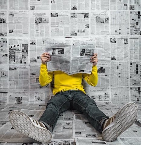 Scelte informate: un giovane legge attentamente un giornale nella stanza piena di testate, evidenziando l'importanza dell'informazione affidabile e l'evitare trappole di notizie false.